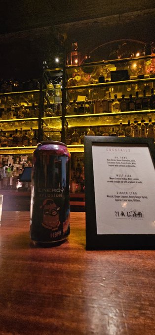 A dimly lit bar scene featuring a dark can of 'Energy Explosion' soda sitting on a wooden counter in front of a backlit shelf of liquor bottles, next to a framed 'Cocktails' menu listing options like 'H.D. Zone', 'West Side', and 'Ginger Lynn' at 'Magma' bar.