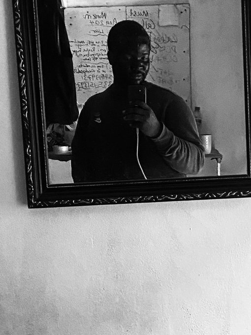 A black-and-white mirror selfie shows a person holding a smartphone in a room with a whiteboard covered in writing.