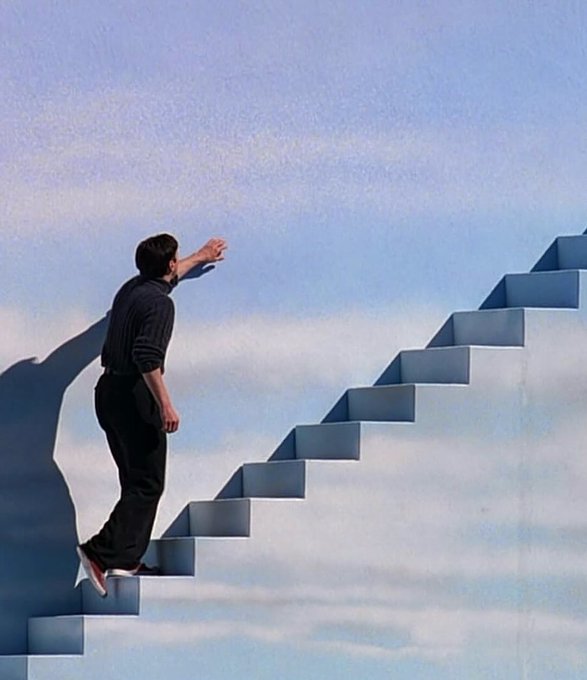 A man in dark clothing climbs a surreal staircase ascending into a soft blue sky, his outstretched hand reaching upward as he steps off the edge into open air.