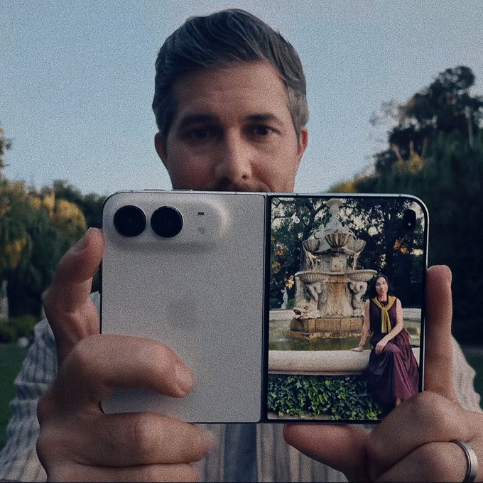 A man holding a white foldable smartphone displaying a photo of a woman in a purple dress sitting by a stone fountain in a garden setting.