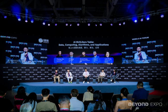 The image shows a group of people sitting around a table, possibly participating in a panel discussion or meeting at an event. The stage has blue lights illuminating the area, creating a professional atmosphere for the gathering. There are several individuals seated on chairs placed around the table, and some attendees can be seen standing nearby. A few handbags are visible among the audience members' belongings.

The image also contains text that reads "@0G_labs 最有意思的地方". This suggests that the event is likely related to a specific company or organization, possibly discussing topics of interest within their industry.