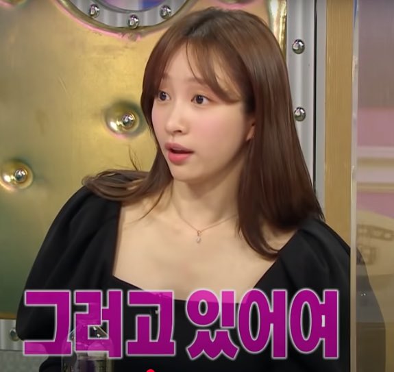 The image shows a young woman with long brown hair wearing a black top and a delicate necklace, appearing mid-speech against a metallic studded backdrop. Pink Korean text overlaid reads "그리고 있어요" ("and is..."), and the post draws an ironic comparison between casually checking for vulnerabilities in Bitcoin hardware wallet code (SeedSigner/Krux) versus the speaker's seemingly offhand conversational tone.