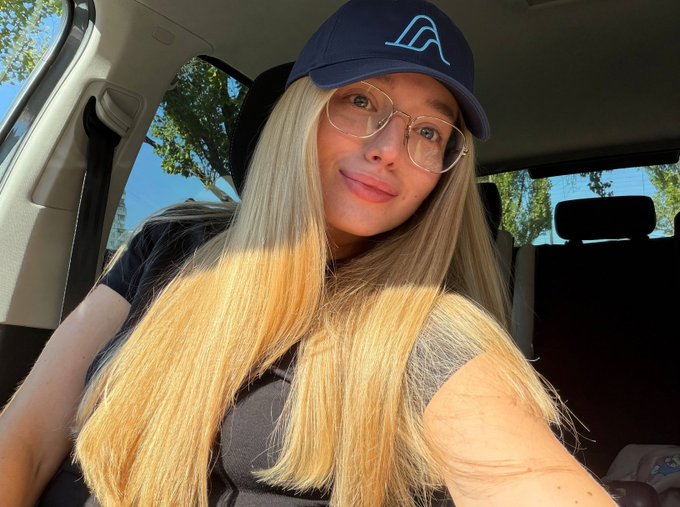 A selfie of a young woman with long blonde hair wearing clear-rimmed glasses and a dark blue baseball cap with a light blue mountain logo, taken inside a car.