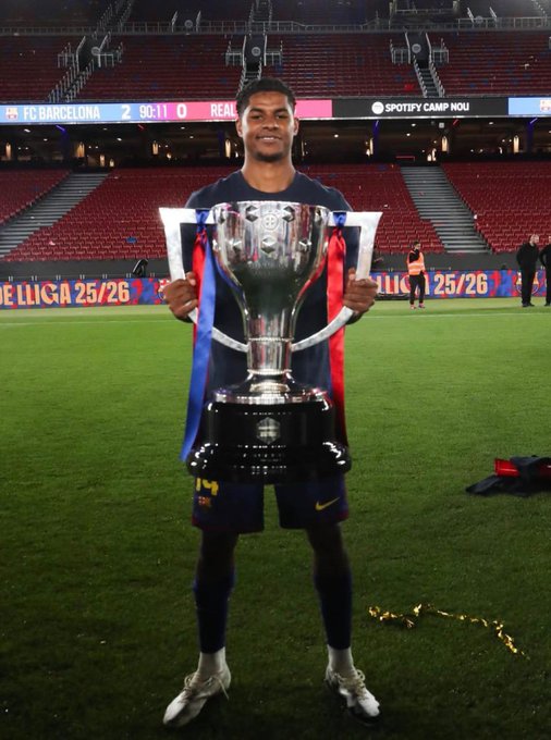 A young soccer player in a blue and red FC Barcelona kit smiling while holding the La Liga trophy on the pitch, with the scoreboard showing 'FC BARCELONA 2 - 0 REAL'.