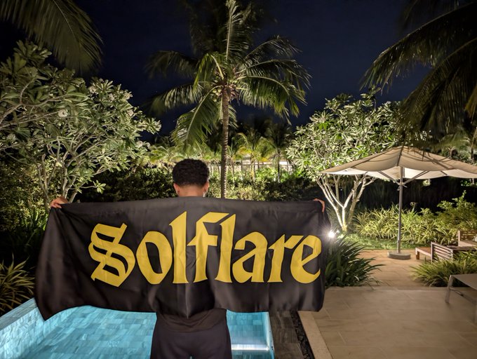 A person holds up a black flag with the word 'Solflare' in yellow gothic lettering, standing by a pool at night surrounded by tropical plants.