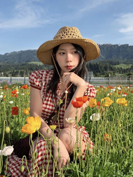A young woman wearing a straw hat and a red-checkered dress sitting in a field of colorful poppies and flowers with mountains and blue sky in the background.