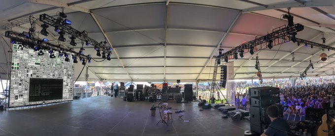 A panoramic view from the stage of a large outdoor music festival tent, showing sound equipment, lighting rigs, and a crowd of attendees in the distance.