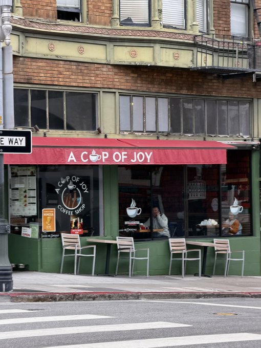 A coffee shop's bright red awning displays "A CUP OF JOY" above large windows, while a "ONE WAY" street sign is visible on the left—showcasing the playful contrast between the cheerful shop signage and the directional traffic sign, perfectly capturing the "seeing signs" theme.
