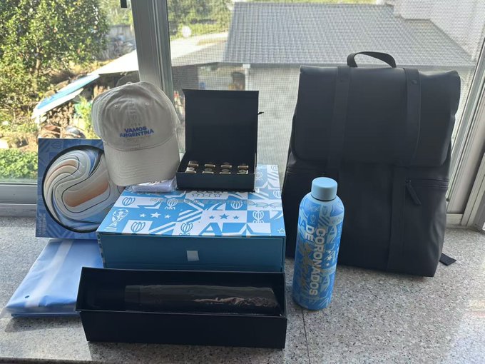 A collection of merchandise and promotional items arranged on a stone surface, including a white 'VALOR ARGENTINA' cap, a black backpack, a blue Bodyarmor water bottle, and decorative boxes.