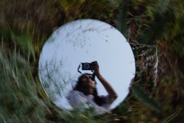 A reflection in a round mirror shows a person holding a camera up to take a photo, framed by tall grass.