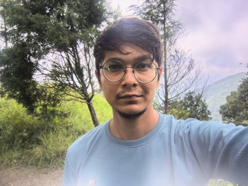 A selfie of a young man with dark hair and round glasses wearing a light blue sweater, standing outdoors with green trees and a hazy mountain landscape behind him.