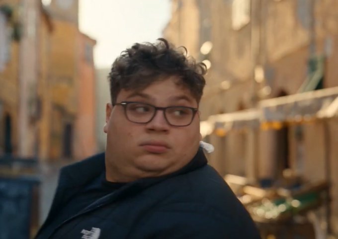 A young man with curly hair and dark-rimmed glasses looks off to the side while standing outdoors in what appears to be a European street setting.