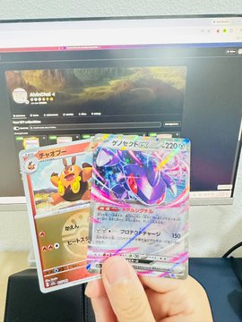 A person holding Japanese Pokémon trading cards, including one featuring Charizard, in front of a laptop screen displaying an auction site.