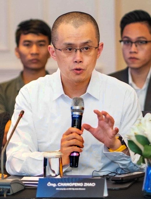 Binance CEO Changpeng Zhao (CZ) speaks into a microphone while gesturing with his hand, seated at a panel with a name placard reading 'Ong. CHANGPENG ZHAO, Chairman and CEO, Binance,' flanked by two other men in the background.