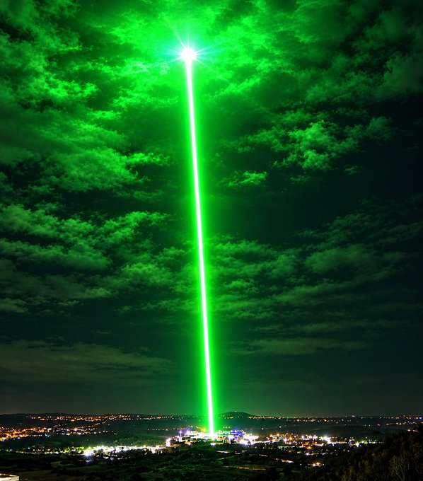 A bright green beam of light shoots up into a cloudy night sky above a city landscape.