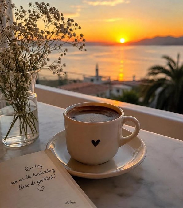 A scenic sunset view over water from a balcony, featuring a coffee cup with a heart on it, a vase of baby's breath flowers, and a handwritten note in Spanish reading 'Que hoy sea un día bendecido y lleno de gratitud!'.