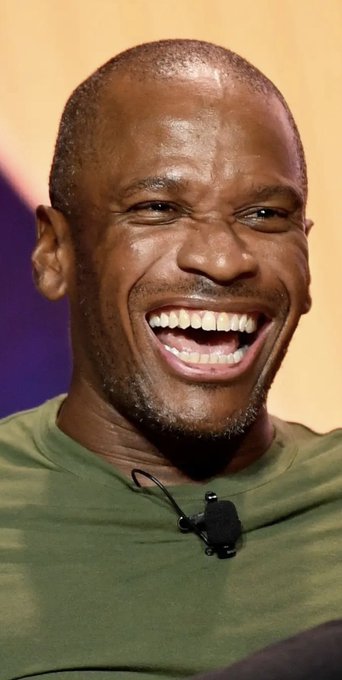 A man with a shaved head wearing an olive-green t-shirt with a small lapel microphone attached laughs heartily, his wide smile and joyful expression suggesting he's enjoying a lighthearted moment during what appears to be a podcast or panel discussion.

The image connects to the post as a visual representation of the person being discussed—"Arthur"—who is being challenged in a trading competition, with the speaker boasting about their profits on $ZEC, $HYPE, $NEAR, and $WLD and looking ahead to outperforming him on $SYN.