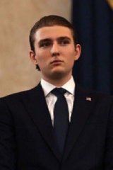 A young man in a dark suit, white shirt, and navy tie with an American flag pin stands against a blue curtain backdrop, looking serious.