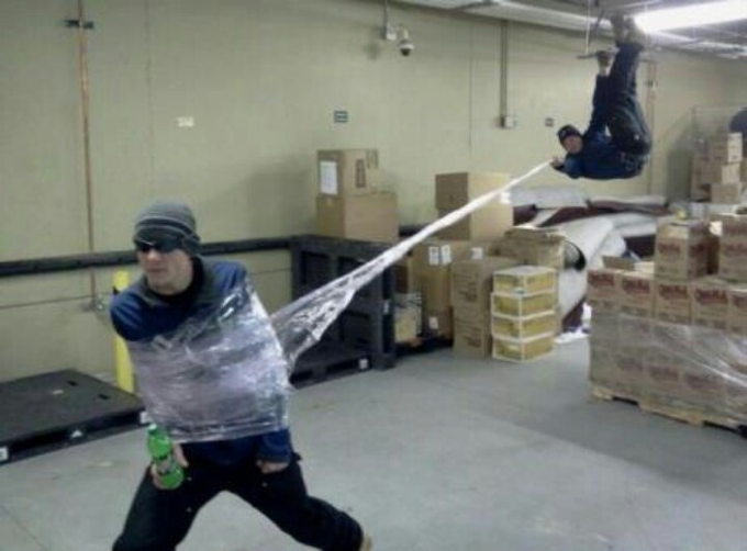 Two men are wrapped in plastic wrap in a warehouse, with one in the background swinging from the ceiling like Spider-Man. The image humorously illustrates the post's caption about being unable to "log off" from the market, as the stocks continue to move—showing the chaos and helplessness traders feel when they can't escape the market's pull.