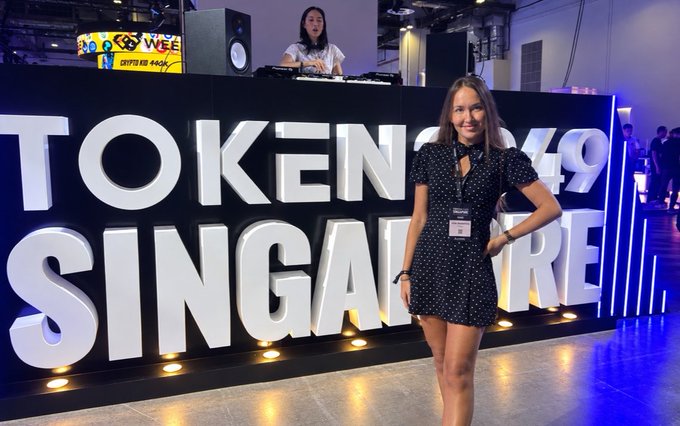 A woman in a black polka-dot dress posing in front of a large 'TOKEN 2049 SINGAPORE' sign at the cryptocurrency conference.