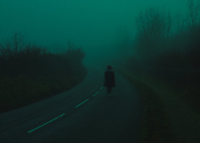 A moody, dark, foggy scene of a lone person walking down an empty road lined with trees, cast in a teal-green hue.