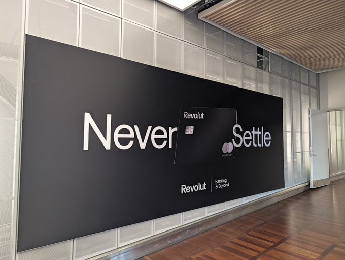 A large black wall display in a modern office lobby reads "Never Settle" with a Revolut credit card propped against it, accompanied by the Revolut logo and the tagline "Banking & Beyond." The image cleverly illustrates the post's warning about the dangers of staying in one place too long by juxtaposing a Revolut advertisement's "Never Settle" slogan—which normally encourages ambition or switching banks—with a cautionary reminder that remaining stationary can leave you vulnerable to threats like robbery.
