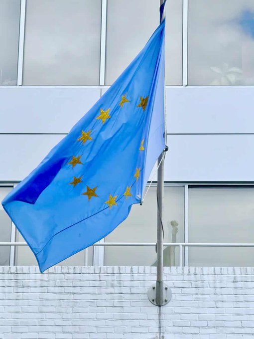 A European Union flag (light blue with gold stars) waves against a clear sky in front of a modern white building, representing regulatory approval. The image complements the article's news about Ripple obtaining full MiCA (Markets in Crypto-Assets) CASP (Crypto-Asset Service Provider) authorization to operate across European markets.