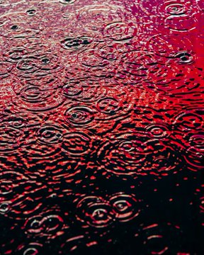 A dark, moody photograph of raindrops creating concentric ripples in a body of water tinted with deep red and magenta hues.