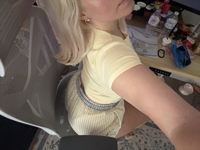 A woman with blonde hair wearing a pale yellow ribbed top and a houndstooth-patterned skirt sits in a mesh office chair, with a cluttered desk behind her holding water bottles, a water bottle, and various items. The messy desk in the background reinforces her caption about not bothering to clean it before a "wowF" event.