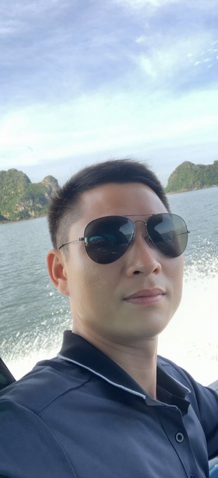 A selfie of a man wearing aviator sunglasses and a dark blue polo shirt, taken on a boat with rocky islands and water visible in the background.