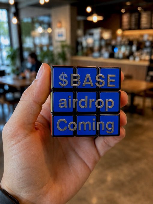 The image shows a person's hand holding up a blue Rubik's cube with the words "Sbase airdrop Coming" written on each side, indicating that the cube will soon be released as an airdrop puzzle game.