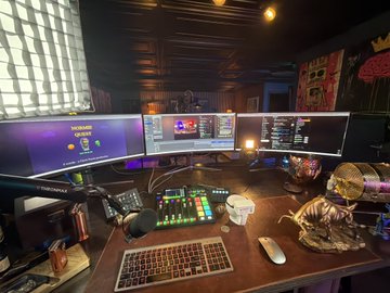 A dimly lit, atmospheric streaming or gaming setup featuring a curved triple-monitor desk arrangement, a backlit keyboard, audio equipment, and a decorative dinosaur skull illuminated by a warm desk lamp.