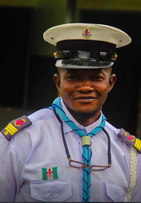 A portrait of a smiling young man in a light blue uniform shirt with shoulder epaulets, a turquoise braided tie, and a white peaked cap with an emblem, with reading glasses hanging from his collar.