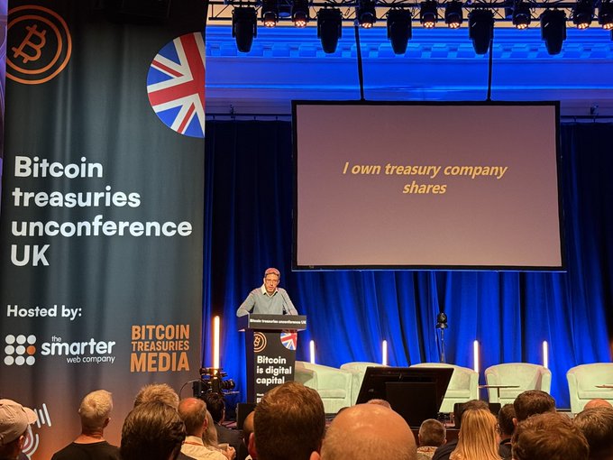A man giving a speech at an event, with a large screen behind him displaying information about Bitcoin and Treasury shares.