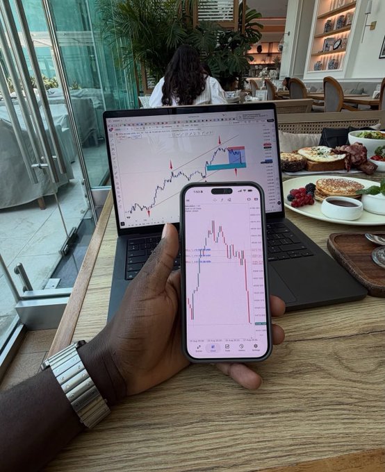 A person's hand holding a smartphone displaying a trading chart in front of a laptop also showing charts, with breakfast on the table at a cafe.