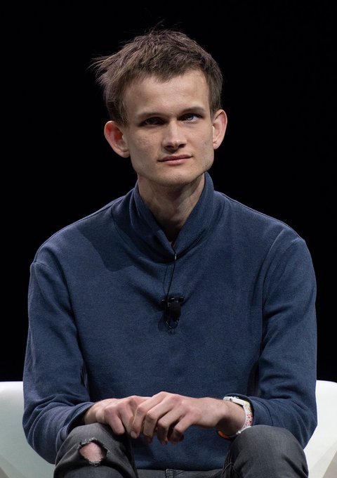A young man with short brown hair, wearing a navy blue mandarin-collar jacket and a wristwatch, sits on a white chair against a black backdrop. The accompanying post humorously addresses "Vitalik" (likely Vitalik Buterin, Ethereum's co-founder) while promoting a crypto trading plan for ETH, but the image itself bears no relation to the text and shows no identifiable text, names, or Ethereum-related imagery.