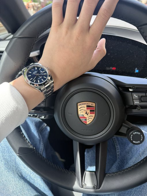 A person's hand rests on a black Porsche steering wheel, showcasing a silver metal watch with a blue dial on their wrist.
