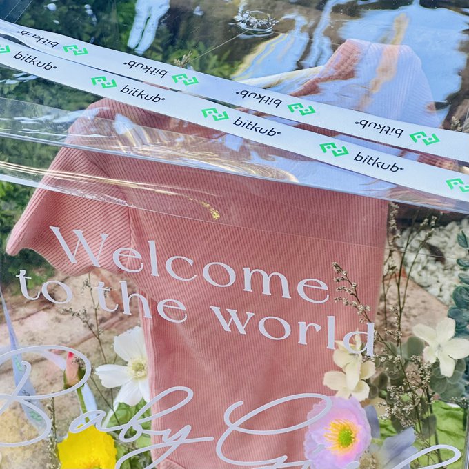 A clear plastic gift bag with 'Welcome to the world' and 'Baby' text printed on it contains a pink garment, sealed with 'bitkub' branded tape.