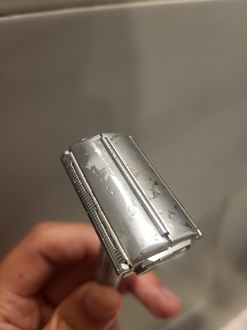 A hand holds up a double-edge safety razor, its chrome head showing smears of shaving cream and small traces of blood, with the background being a tiled shower wall. The image humorously illustrates the post's contrast — while a careless developer might skim funds from a treasury, the dedicated developer endures the painful, nick-prone ritual of traditional wet shaving.