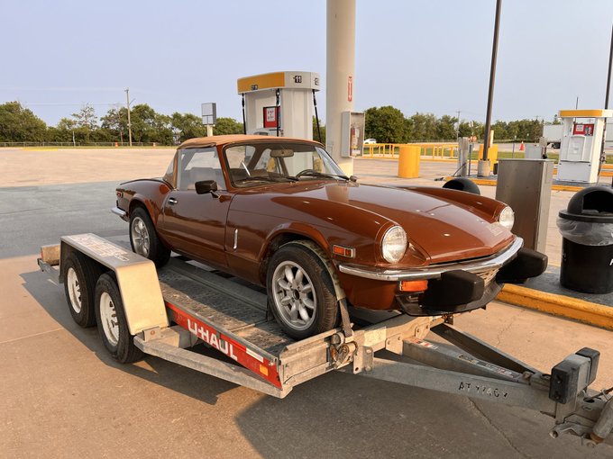 A bronze Triumph Spitfire convertible sits loaded on a U-Haul car trailer at a Shell gas station, marking the "grail" find for its new owner.
