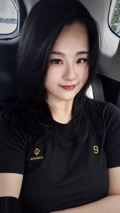 A young woman wearing a black Binance t-shirt (featuring the yellow Binance logo and the number "9") smiles at the camera from inside a vehicle, likely posting in response to a Binance fan art contest as a "#BinancePeripheryAmbassador."