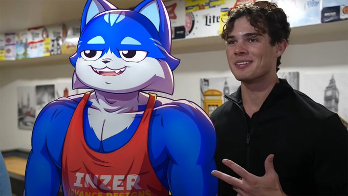 A young man with curly brown hair stands next to an animated blue wolf character wearing an orange Inzer tank top, appearing to be in a kitchen setting.