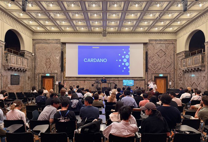 A packed auditorium at the University of Zurich Summer School features a large "CARDANO" branded presentation slide on screen, as the CEO speaks to an engaged audience about building within the Cardano ecosystem—the shared stage that united developers during this first-time company appearance at the event.
