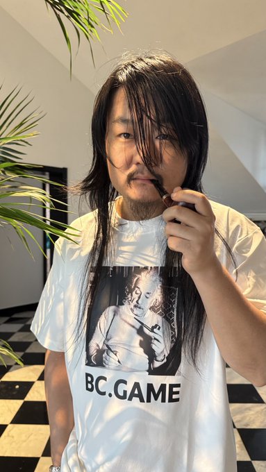 The image features a man wearing a white t-shirt with a picture of a person on it that says "BC Game" written in black letters. The man has long hair, and he's holding his hair up with one hand while using another hand to hold a toothbrush or comb. There is also a potted plant visible in the background.