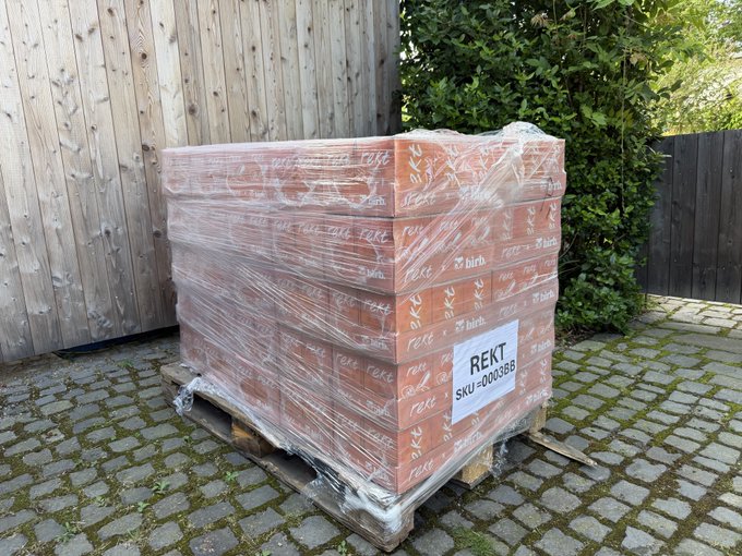 A pallet of bricks wrapped in plastic sits on a paved driveway, with a label reading "REKT" and "SKU-0003BB" — a pun playing on the text "Get Rekt," as bricks would symbolize being "rekt" (i.e., financially ruined) in internet slang.