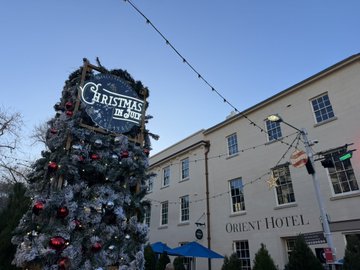 An outdoor Christmas tree decorated with silver and red ornaments, bearing a 'CHRISTMAS IN THE' sign, set against a blue sky in front of the historic ORIENT HOTEL.