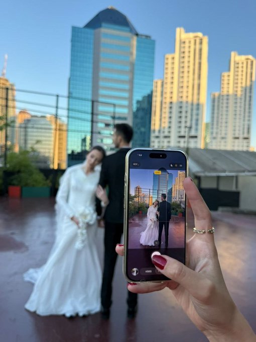 A wedding photo of a bride and groom being taken with a smartphone, with a modern city skyline in the background.