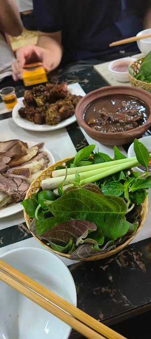 A table spread with various Vietnamese dishes including fresh herbs, sliced meat, fried food, and a brown dipping sauce, with chopsticks and a person's hands visible.