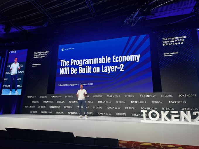 A man speaking on stage at the Token2049 event in Singapore (October 2026), with a large blue screen behind him displaying the title 'The Programmable Economy Will Be Built on Layer-2' by Steven Goldfeder of Offchain Labs.