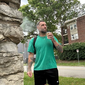 A man with tattoos wearing a green t-shirt and black shorts standing outdoors near a stone wall, holding something while exhaling visible vapor or smoke.